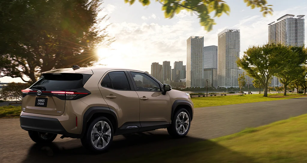 公園沿いの道にいるベージュのヤリス クロスを後ろ斜めから見た外観。テールランプと背面のYARIS CROSSの文字が見える