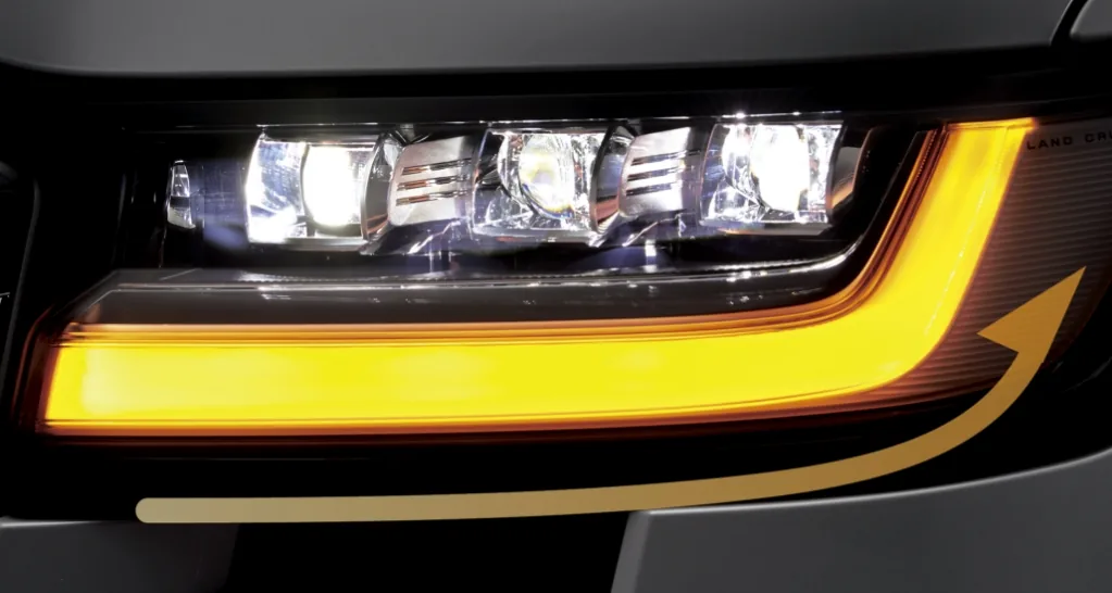 ランドクルーザー300のLED式ヘッドランプのアップ。上に3つの光源が並び、その下に黄色い帯状のランプが横に光り、金色の矢印の図が重なっている