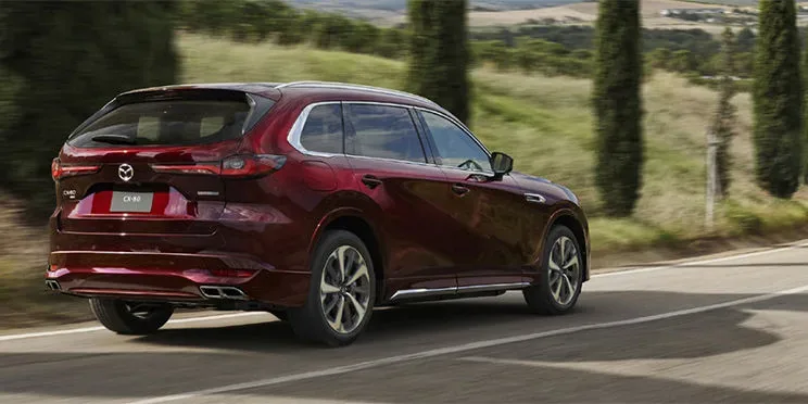 糸杉の並ぶ道を走る赤いCX-80を左斜め後ろから見た様子。横長のテールランプと、左右に分かれたマフラーの出口が見える