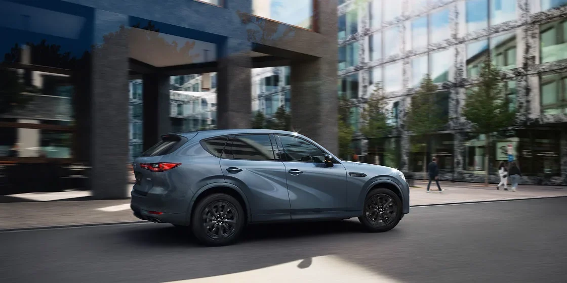 ビルの前の道を走る青灰色のCX-60を、斜め後ろ寄りから見た様子。黒いホイールが見える