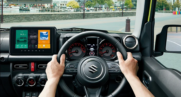ジムニーの運転席まわり。ステアリングを両手で握った視点で、中央のディスプレイオーディオとメーターが見える公式写真